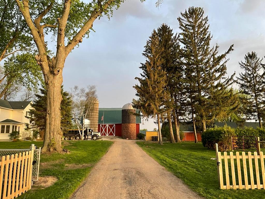Scenic rural driveway leading to a traditional farmstead, featuring a red barn, silos, and lush greenery, framed by tall trees and a white picket fence. Ideal for showcasing farm life and countryside aesthetics.