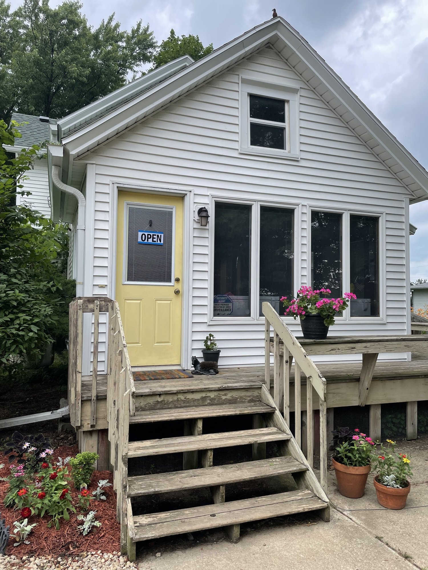 Charming white house with yellow door and "Open" sign, featuring a wooden front porch with steps, surrounded by colorful flower pots and vibrant landscaping. Ideal for welcoming visitors.