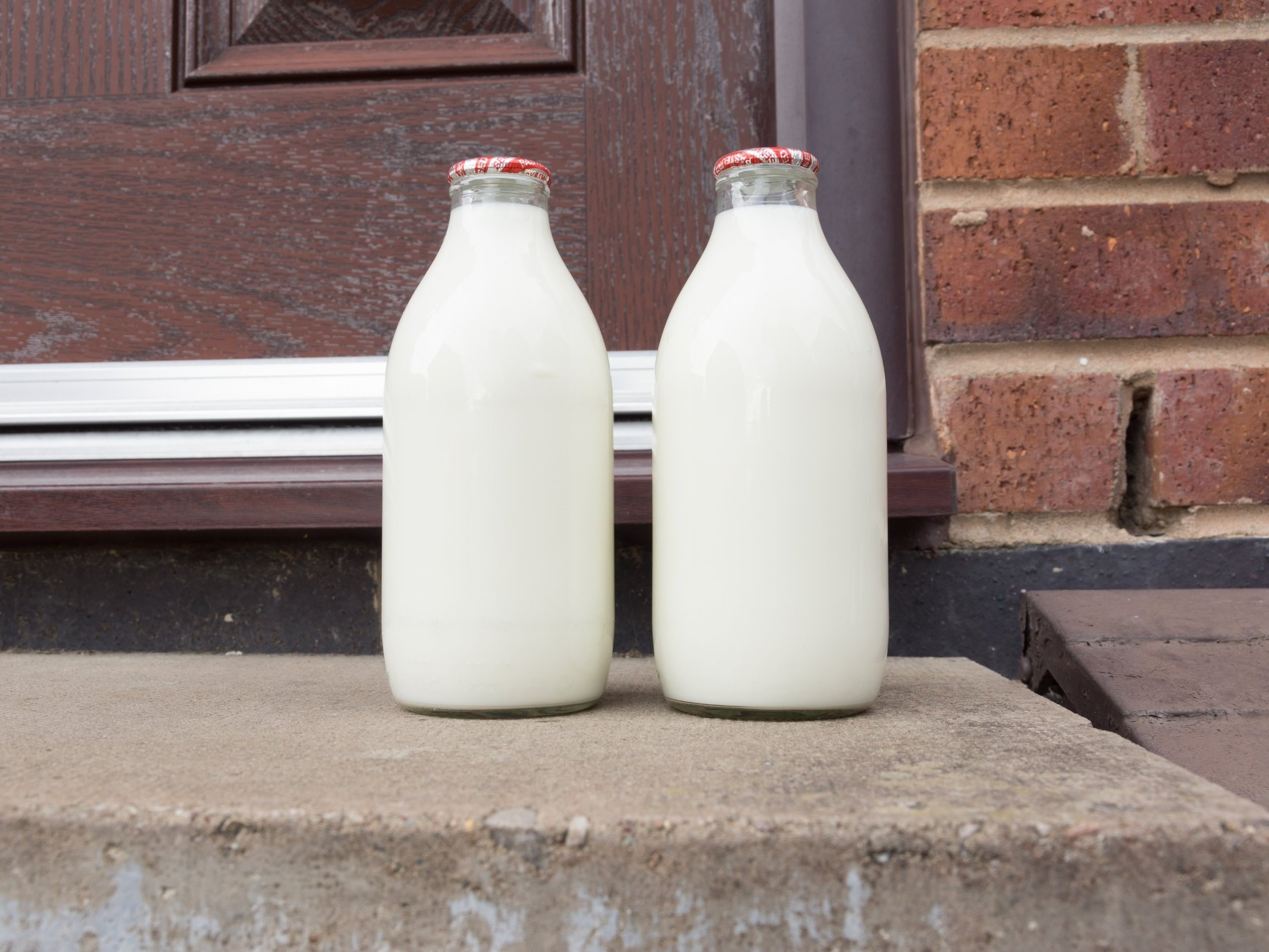 Milk_bottles_on_doorstep (1) Two glass bottles of fresh milk sit on a concrete step in front of a wooden door, showcasing a rustic, home-delivery vibe.