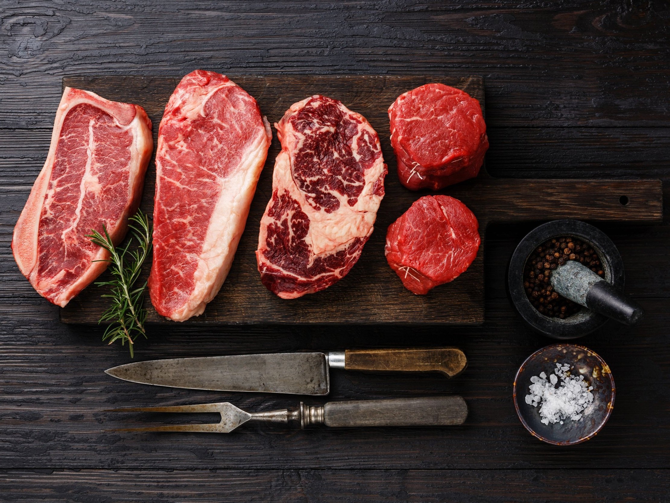 beef_nystrip_ribeye_tenderloin_3 (6) Assortment of fresh raw steaks including ribeye, sirloin, and tenderloin on a wooden cutting board, accompanied by herbs, a mortar and pestle with spices, and a bowl of sea salt, ideal for grilling or cooking enthusiasts.