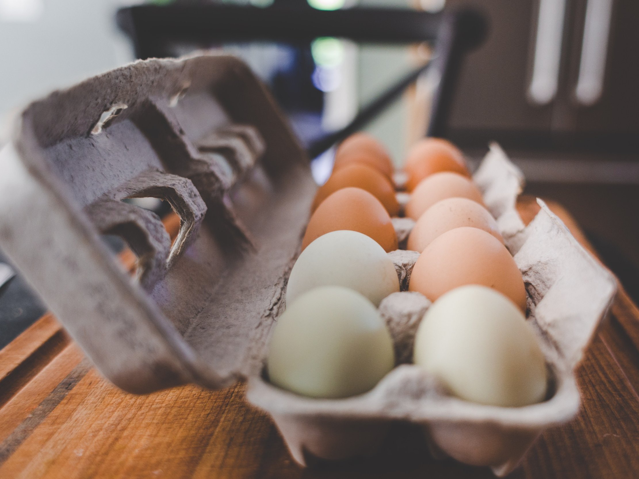 egg_carton_oncounter Carton of eggs featuring a variety of colors, including brown and green, arranged neatly on a wooden surface, with a blurred kitchen background. Ideal for showcasing fresh, organic eggs for cooking and baking.