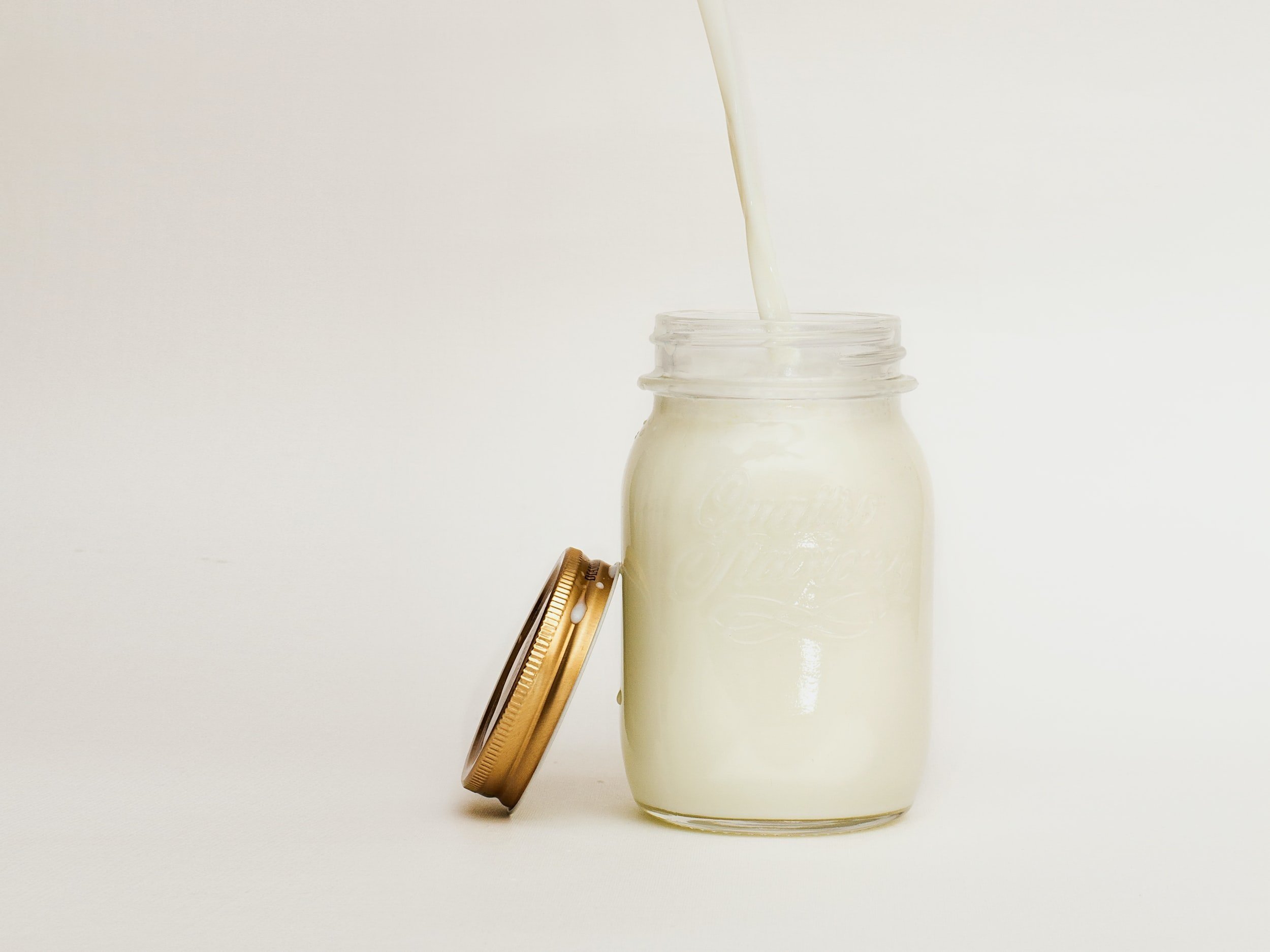 Pouring fresh milk into a glass jar with a golden lid, set against a plain white background.